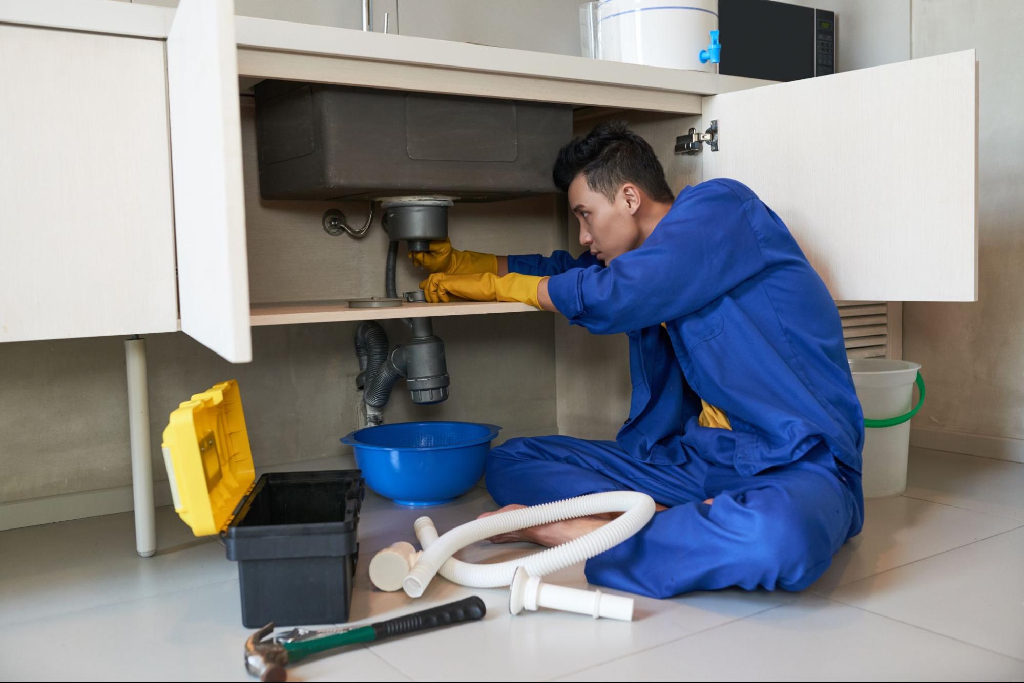Technician in blue overalls repairing pipes under a kitchen sink.